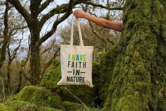 Faith In Nature tote bag held beside mossy trees in a forest