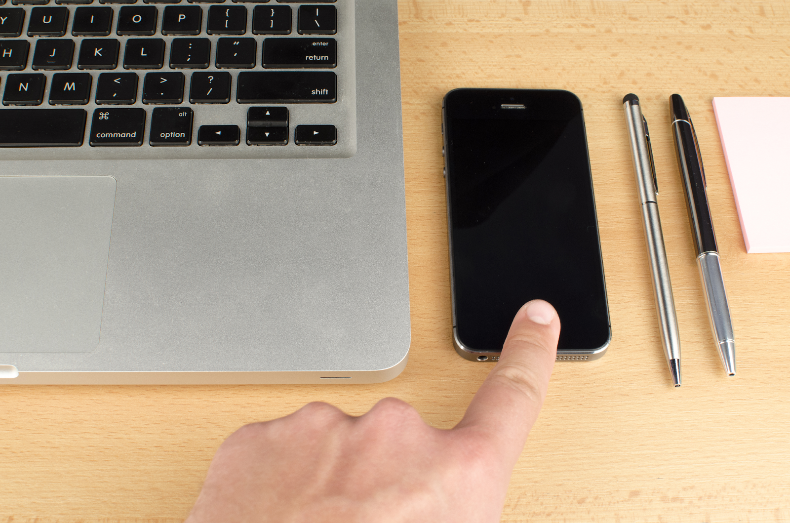 A finger hovering above a smartphone between a laptop and pens