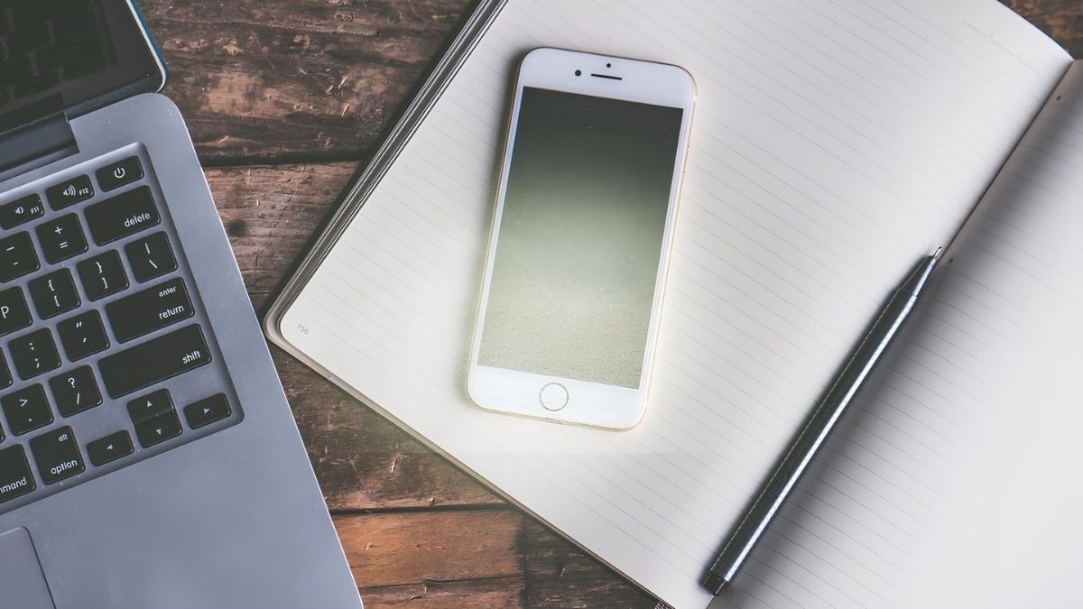 A white smartphone resting on an open notebook beside a pen and laptop
