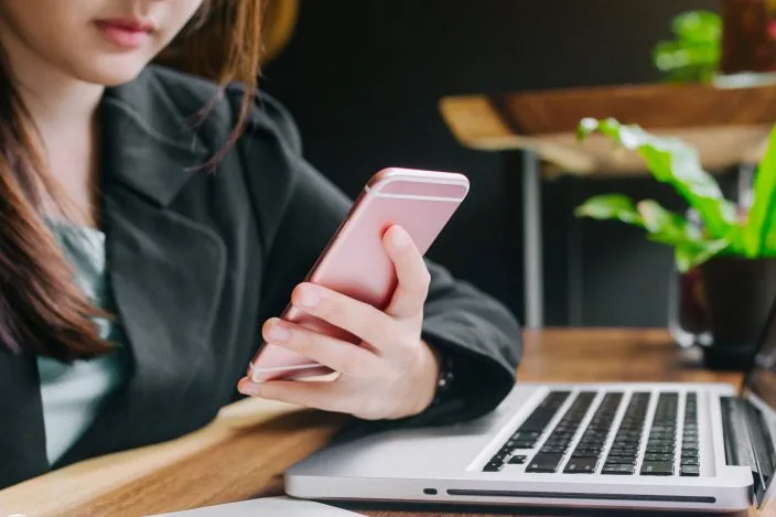 A person holding a smartphone beside an open laptop at a desk